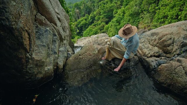 Middle-aged woman collecting pure water from natural rock pool above tropical valley, clean stream and lush green mountains creating a calm and eco travel scene
