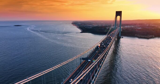 Flight over the multi-lane roads with hectic traffic on the Verrazano- Narrows Bridge in New York City, USA. Amazing orange sky at sunset in the backdrop.