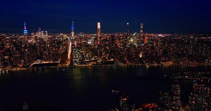 Boat moves by the dark East River waterscape at night. Scenery of iconic Manhattan across the river from drone. NYC, USA.