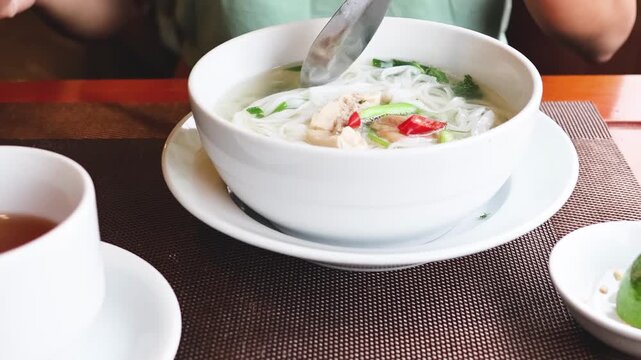 Close up of person eating pho noodles with chopsticks and spoon in a white bowl with tea.