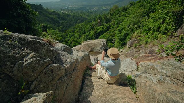 Young woman sitting on rocky ledge above lush tropical valley, capturing scenic mountain landscape on smartphone during peaceful island travel moment