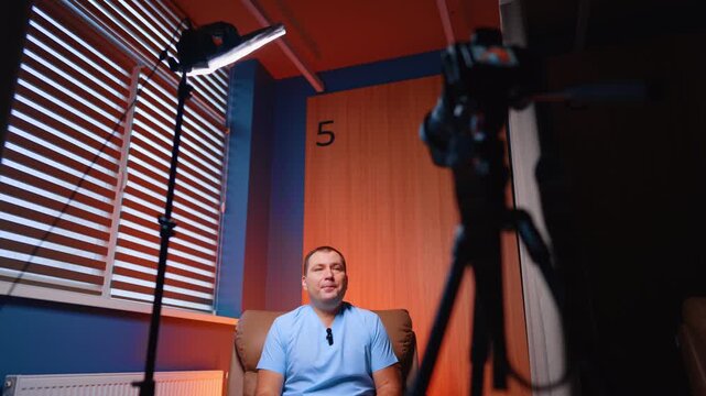 Positive Caucasian man in blue uniform sits in an armchair in the light of soffit. Medic speaks to camera directed on him. Blog concept.
