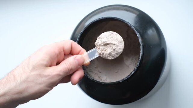 Male hand holds measuring spoon with brown protein on white background top view.