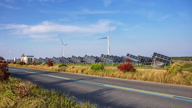 Time-lapse of coastal hybrid renewable energy farm with tracking type solar panels, wind turbines, and scenic tourist road under blue sky at dusk 