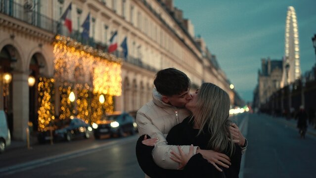 A romantic couple is kissing on a street in Paris France at dusk with city lights and a Ferris wheel in the background. This image can be used for travel romance and European destination concepts.