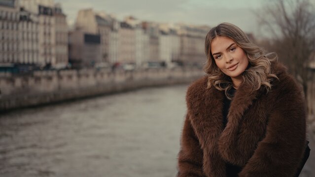 A blonde woman poses in a brown fur coat with the Seine River and city buildings in the background in Paris France. This image is suitable for fashion lifestyle and travel content.