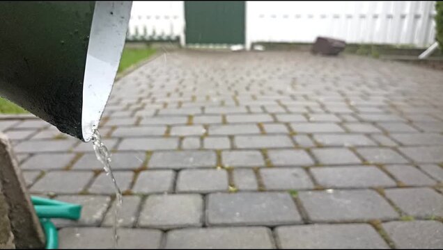 Close-up video of rainwater flowing from a metal gutter pipe onto wet pavement during rainy weather near a residential driveway.
