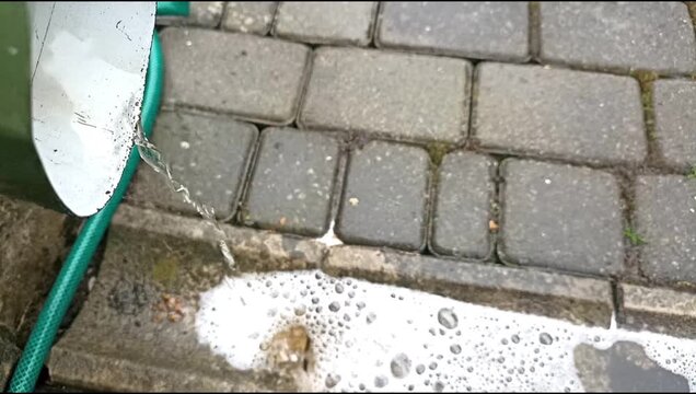 Close-up video of rainwater flowing from a metal gutter pipe onto wet pavement during rainy weather near a residential driveway.