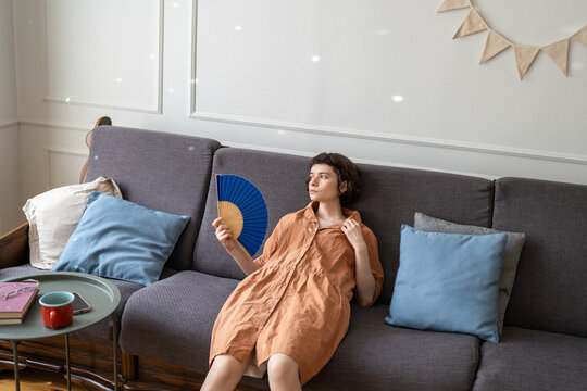 Summer heat and no air conditioning, tired woman lying back on couch waving hand fan feeling uncomfortable and exhausted from overheating, female with paper fan suffers from hot weather at home.