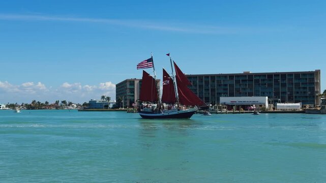 Side view of a beautiful sailboat with red sails and American flag sailing on turquoise water heading out to sea in front of waterfront buildings