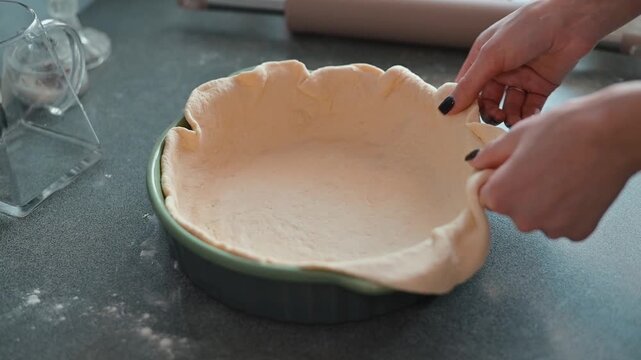A person carefully shapes and molds pastry dough into a pie dish in a kitchen.