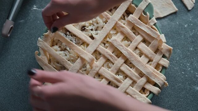 A person prepares a pie by placing a lattice top over the filling in a baking dish. The individual is focused on creating an even pattern with their hands