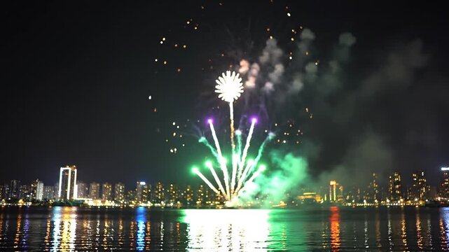Colorful fireworks exploding over city skyline at night reflecting in water during celebration.