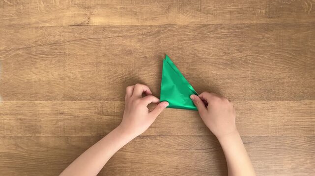 child hands folding green triangular origami paper into smaller triangle on wooden table slow motion 4K