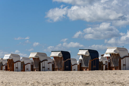 View of traditional hooded beach chairs lined up on the sandy shore of Strand Schillig under a bright blue sky with fluffy white clouds, Wangerland, Niedersachsen, Germany.