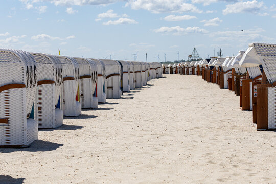 View of rows of traditional white wicker hooded beach chairs on the sandy Strand Schillig under a bright sky with wind turbines in the distance Wangerland, Niedersachsen, Germany.