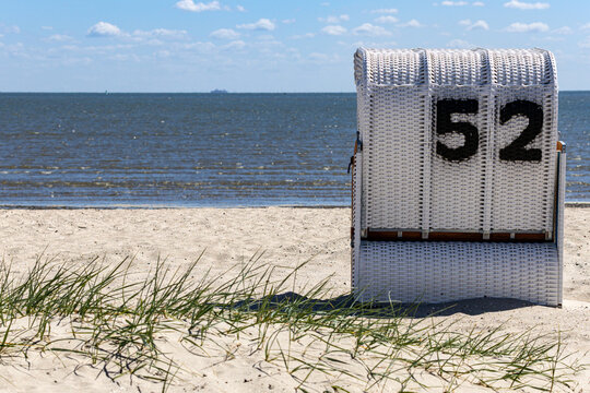 View of a white wicker beach chair with number 52 on the sandy Strand Schillig beach overlooking the North Sea under a blue sky in Wangerland, Niedersachsen, Germany.