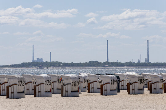 View of Strand Schillig with rows of numbered white hooded beach chairs on the sand and industrial chimneys on the horizon across the water in Wangerland, Niedersachsen, Germany.