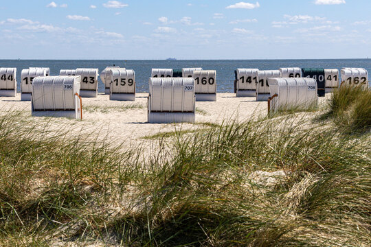 View of Strand Schillig with numbered white hooded beach chairs on the sand and green dunes under a blue sky in Wangerland, Niedersachsen, Germany.