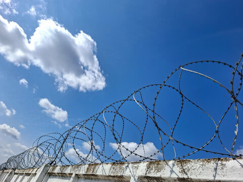 Coiled razor wire fence on concrete wall with blue sky and clouds above, representing security and restriction in urban area