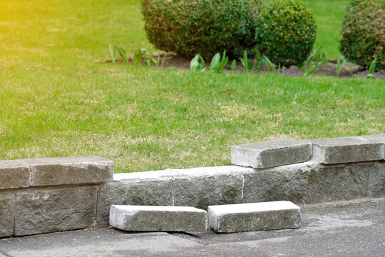 Damaged low retaining wall with fallen bricks, need for masonry repair in urban landscaping. Crumbling stone border of green garden. Damage of low grey stone retaining wall or garden border