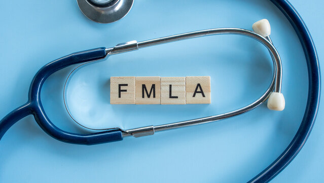Medical stethoscope framing wooden letter tiles that spell out FMLA acronym representing Family and Medical Leave Act on blue surface