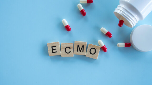 Wooden blocks spelling the acronym ECMO next to an open pill bottle with red and white capsules spilled on a blue surface