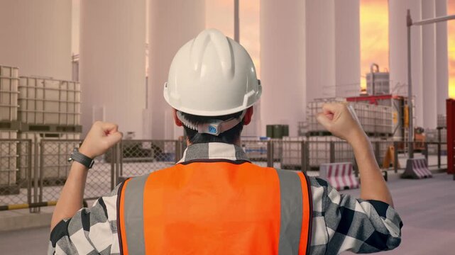 Close Up Back View Of A Male Engineer With Safety Helmet Raising His Hands Celebrating While Working at Storage Facility with IBC Totes, Large Silos at Sunset