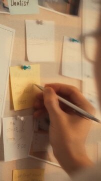 In cozy workspace, person pins pictures and documents to board while scribbling ideas on a sticky note, deep in a brainstorming session. The setup blends creativity and organization as thoughts form