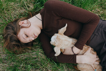 Fototapeta premium A woman lies on grass in a brown sweater, holding a gentle goat kid. The scene conveys a calm countryside lifestyle, natural bond, and tender animal companionship outdoors.
