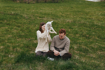 Fototapeta premium A couple sits on grass with a playful kid goat nearby, sharing a tender outdoor moment in a natural meadow, cozy sweaters, relaxed mood, and rustic countryside ambiance.