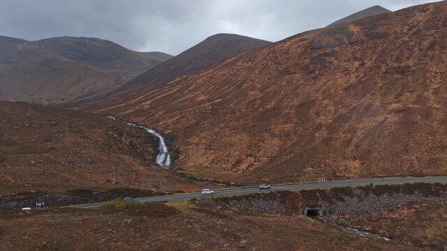 Stunning view of eas a' bhradain waterfall and a scenic road with cars driving through the majestic scottish highlands on a cloudy day