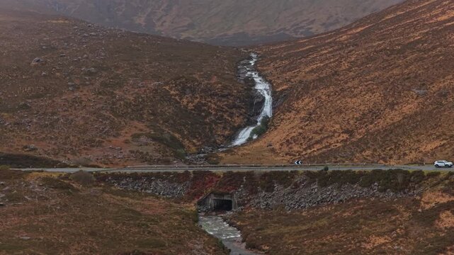 Aerial view of the eas a' bhradain waterfall carving a path through the rusty colored scottish highlands as cars drive on a nearby road