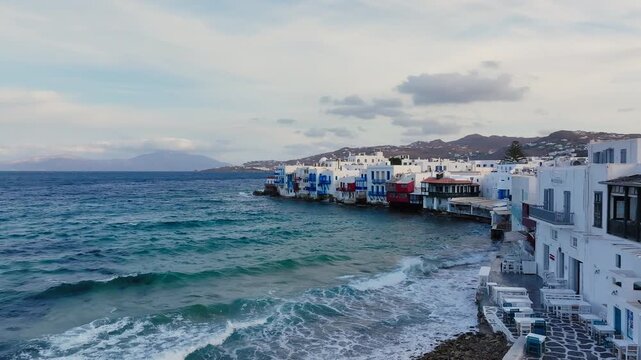 Panoramic drone footage approaching Little Venice, Mykonos island, Greece, on a windy day, colourful neoclassical houses and balconies perched directly above a wavy Aegean Sea