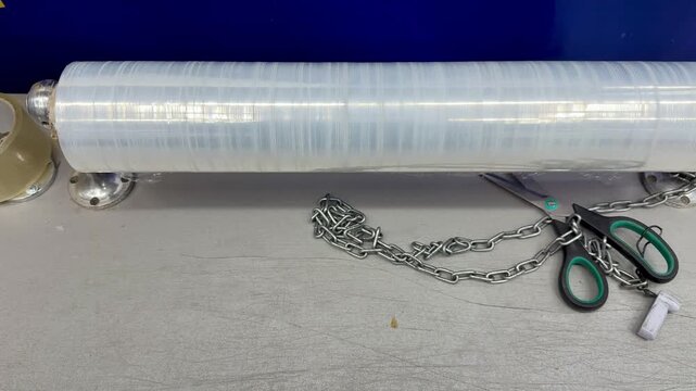 Clear stretch film roll on counter, packaging station with scissors, tape dispenser and metal chain, textured tabletop background