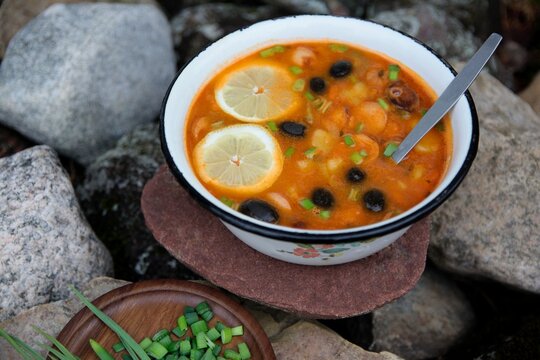 Homemade traditional Eastern European solyanka soup in enamel bowl with spoon inside , outdoor meal on natural stones background, side view