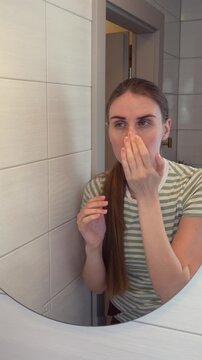 Young woman looking at facial skin in bathroom mirror while checking skin condition and appearance. Concept of skincare routine, beauty care, self examination and dermatology concern.