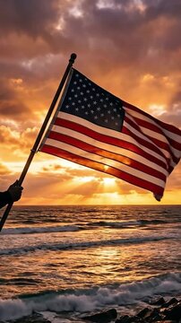 Male hand holding a large American flag above ocean waves during dramatic sunset with glowing clouds and golden sun rays, creating a powerful patriotic and cinematic atmosphere.
