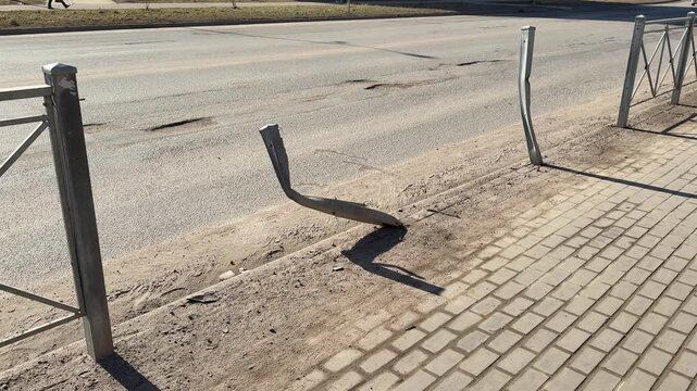 Bent fence post on sunny highway roadside twisted metal leaning over paved sidewalk, gravel scatter and scrape marks from impact