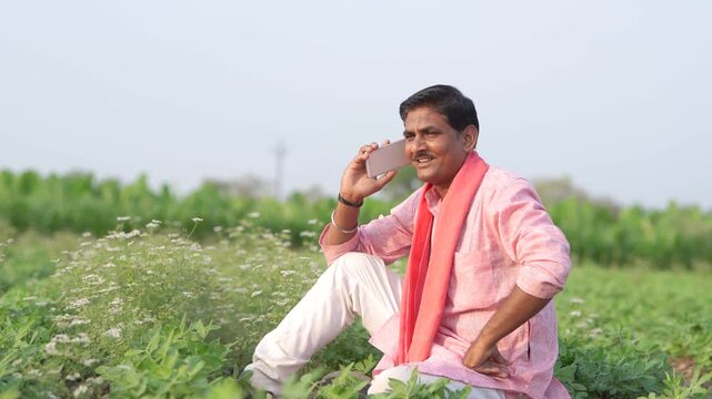 young indian farmer using smartphone sitting at green agricultural field