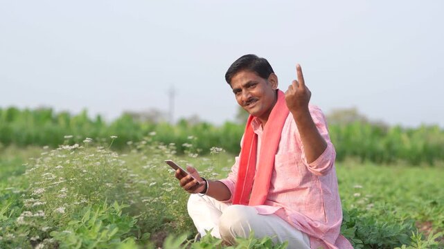 young indian farmer using smartphone sitting at green agricultural field