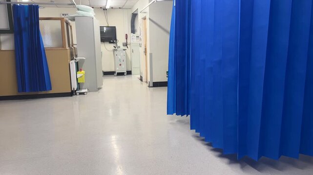 Panning shot of empty cubicles in hospital ward. Blue partitioning curtains hanging open as there are no patients at this time.