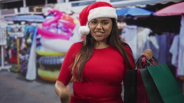 Woman in santa hat and red top holds multiple shopping bags and blows a kiss with her right hand on street market; joyful holiday cheer.