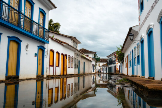 View of historic colonial buildings with colorful doors and windows reflected in a flooded cobblestone street under a cloudy sky in Paraty, State of Rio de Janeiro, Brazil.
