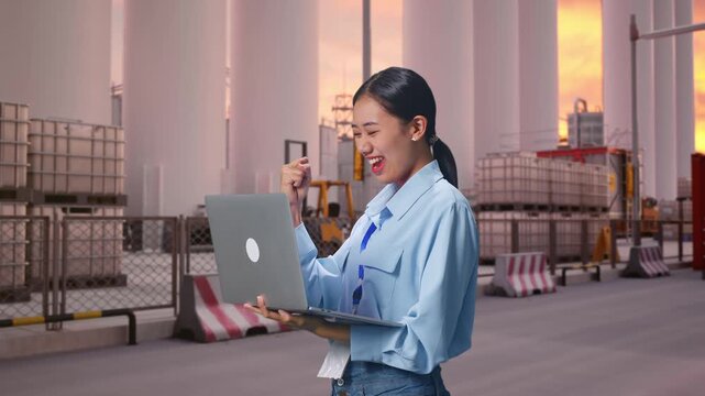 Side View Of Asian Female With Her Laptop at Storage Facility with IBC Totes, Large Silos at Sunset, She Raises Her Fist Up With Screaming Goal After Typed On The Laptop