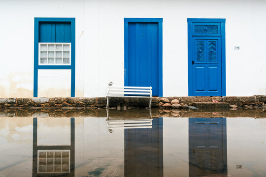 View of colonial architecture with vibrant blue doors and a white bench reflected in the flooded street of the historic center in Paraty, State of Rio de Janeiro, Brazil.