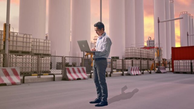 Full Body Side View Of An Asian Male Professional Worker Standing With His Laptop at Storage Facility with IBC Totes, Large Silos at Sunset, Typing On His Laptop'S Keybaord With Meditation