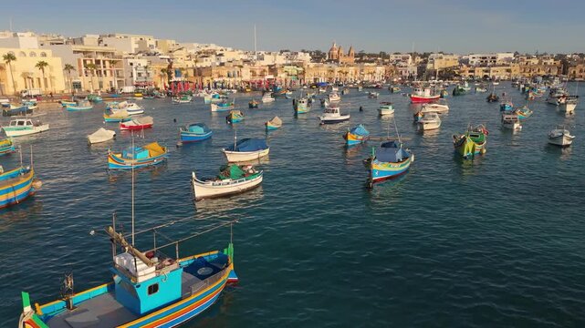 Colorful luzzu boats anchor in bay of Marsaskala, Malta in this aerial 4K. City buildings border busy port in Marsaskala, Malta. Traditional maritime fleet floats in blue sea of Marsaskala, Malta