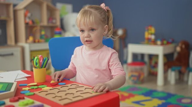 Little blonde girl placing a blue wooden shape into a pegboard puzzle on a red table in a preschool classroom with toys, shelves and a play rug; focused concentration.