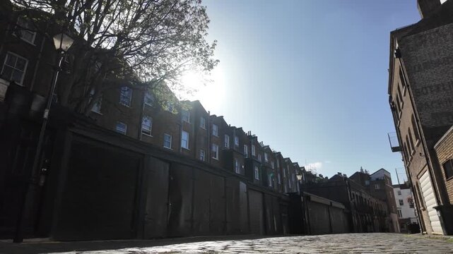 Low angle dolly shot moving backwards along an old empty cobblestone street in london, with traditional brick buildings, sun flare and a clear blue sky on a sunny day